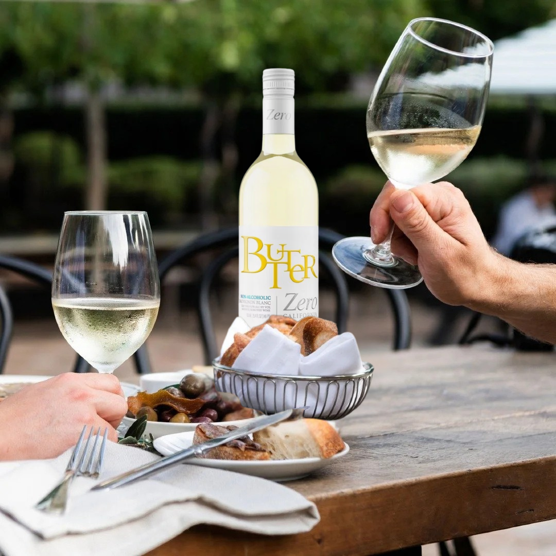 A bottle of white wine with a large yellow 'Butter Zero' label stands on a wooden outdoor table, surrounded by two wine glasses being held by hands, bread in a wire basket, and plates with food. The background shows blurred patio chairs and greenery.