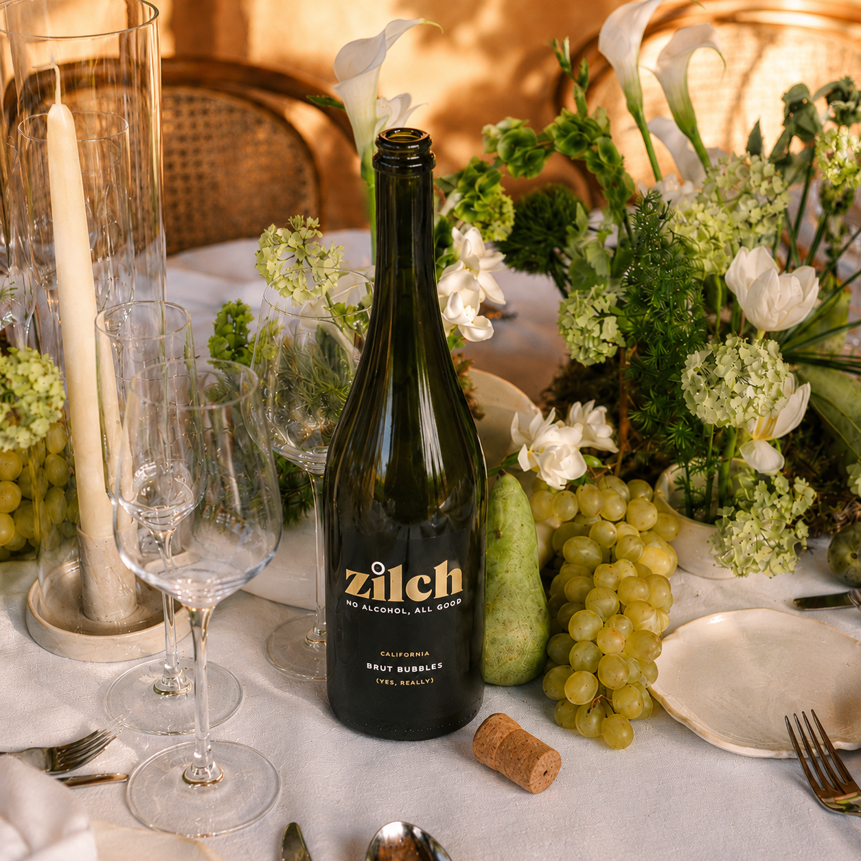 A bottle of Zilch No Alcohol Allergy Brut Bubble wine with a cork, styled on a white tablecloth with wine glasses, candles, and green grapes surrounded by flowers.