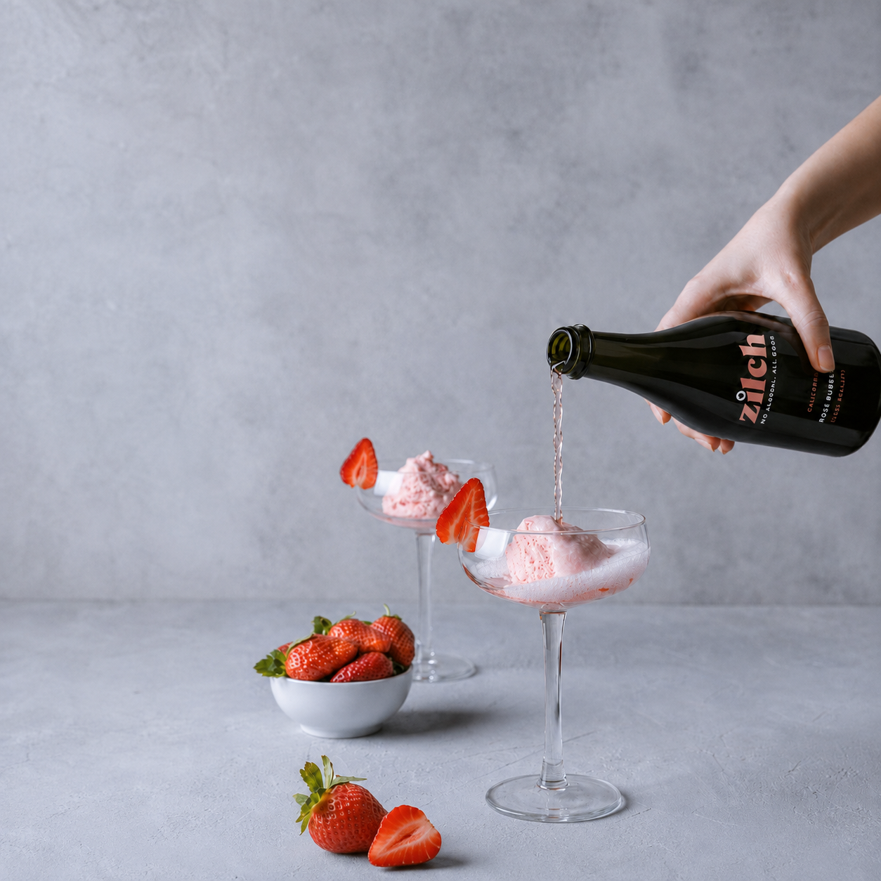 A hand pours a pink strawberry drink from a dark glass bottle labeled 'zich' into two coupe glasses garnished with strawberries, set on a light gray surface against a textured gray background.