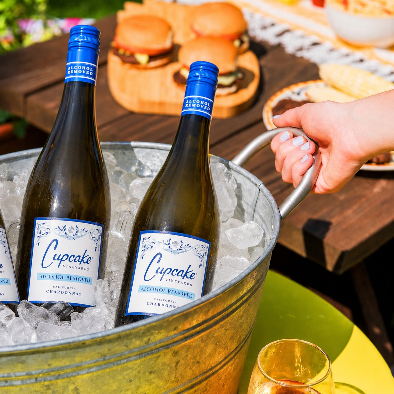 Two bottles of Cupcake Vineyards Alcohol Resistant Chardonnay in a galvanized metal bucket filled with ice, with a hand holding the bucket handle in an outdoor patio setting.