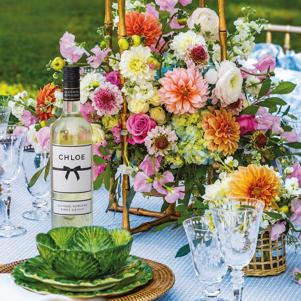 A bottle of Chloe Prosecco Non-Alcoholic Flavour Enhancer stands on a blue tablecloth surrounded by a colorful floral centerpiece, clear drinking glasses, and green ceramic plates in an outdoor dining setting.