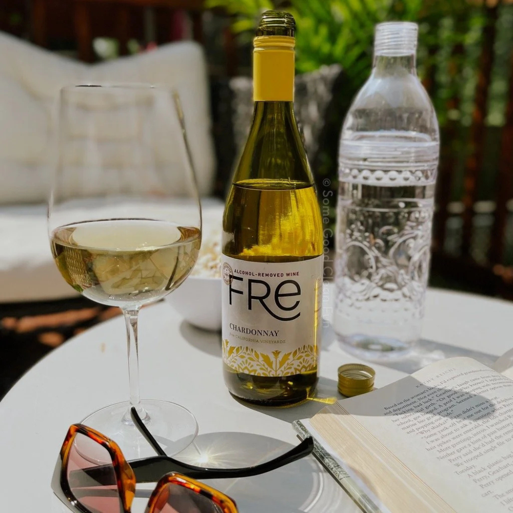 Wine bottle, glass of wine, water bottle, sunglasses, and book on a table outdoors.