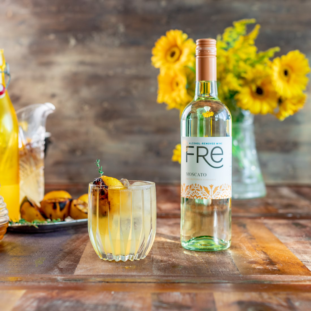 Bottle of Fre Moscato wine on a wooden table with a glass of yellow liquid and flowers in the background.
