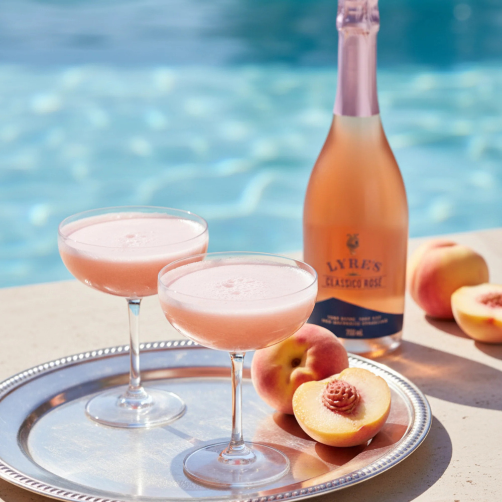 Two glasses of pink cocktail with Lyres Classic Rosé bottle and peaches on a tray by a pool.