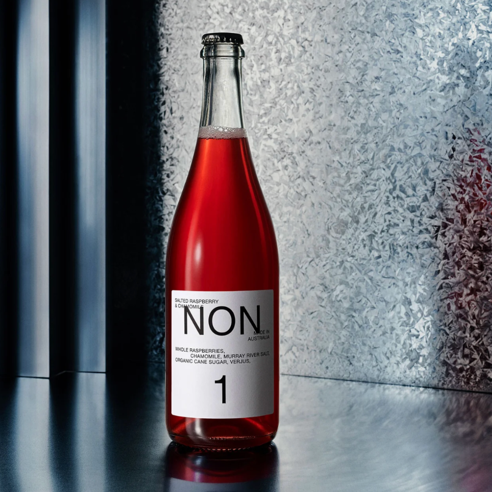 A glass bottle of NON 1 Raspberry Sparkling Wine with a white label, filled with vibrant pinkish-red liquid, set against a textured frosted glass background on a dark reflective surface.