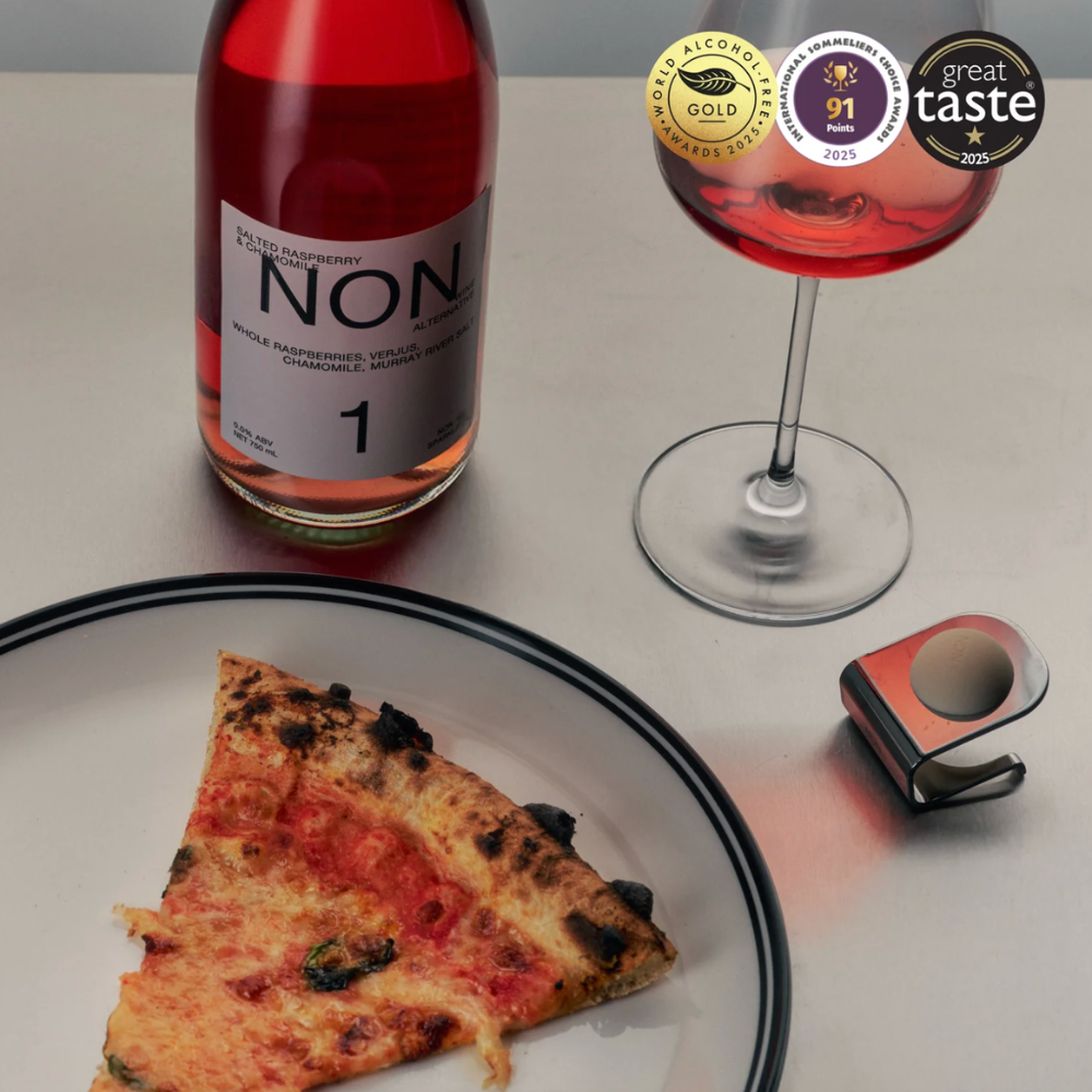 A bottle of NON. single varietal white wine with a slice of pizza on a plate and a wine glass filled with red wine, alongside a small red and black accessory, set on a light gray surface with award badges at the top right.