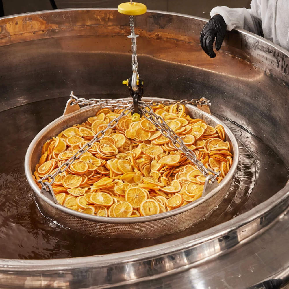 A large metal vat filled with sliced orange peels or dried citrus pieces, suspended in a large pot of water by a chain and pulley system. A gloved hand is visible at the edge of the vat.