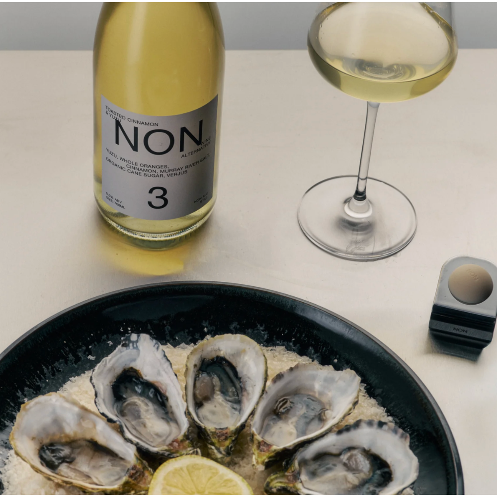 A bottle of Non Alcoholic White Wine with a glass of the pale wine, and a black plate of raw oysters on ice with a lemon wedge, arranged on a light beige tabletop.