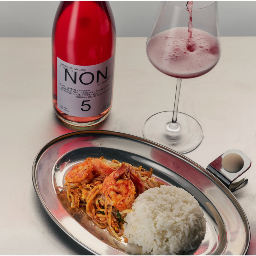 A bottle of NON. Non-alcoholic wine with a pinkish-red liquid and a glass being filled, set on a tabletop next to a plated meal with shrimp pasta, white rice, and a small condiment dish.