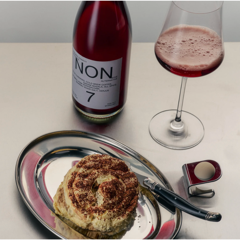 A bottle of red wine with a large white label reading 'NON' stands beside a filled wine glass on a light tabletop. In the foreground, a silver platter holds a pastry topped with brown sugar or cocoa powder, with a knife and a small red-and-white container nearby.