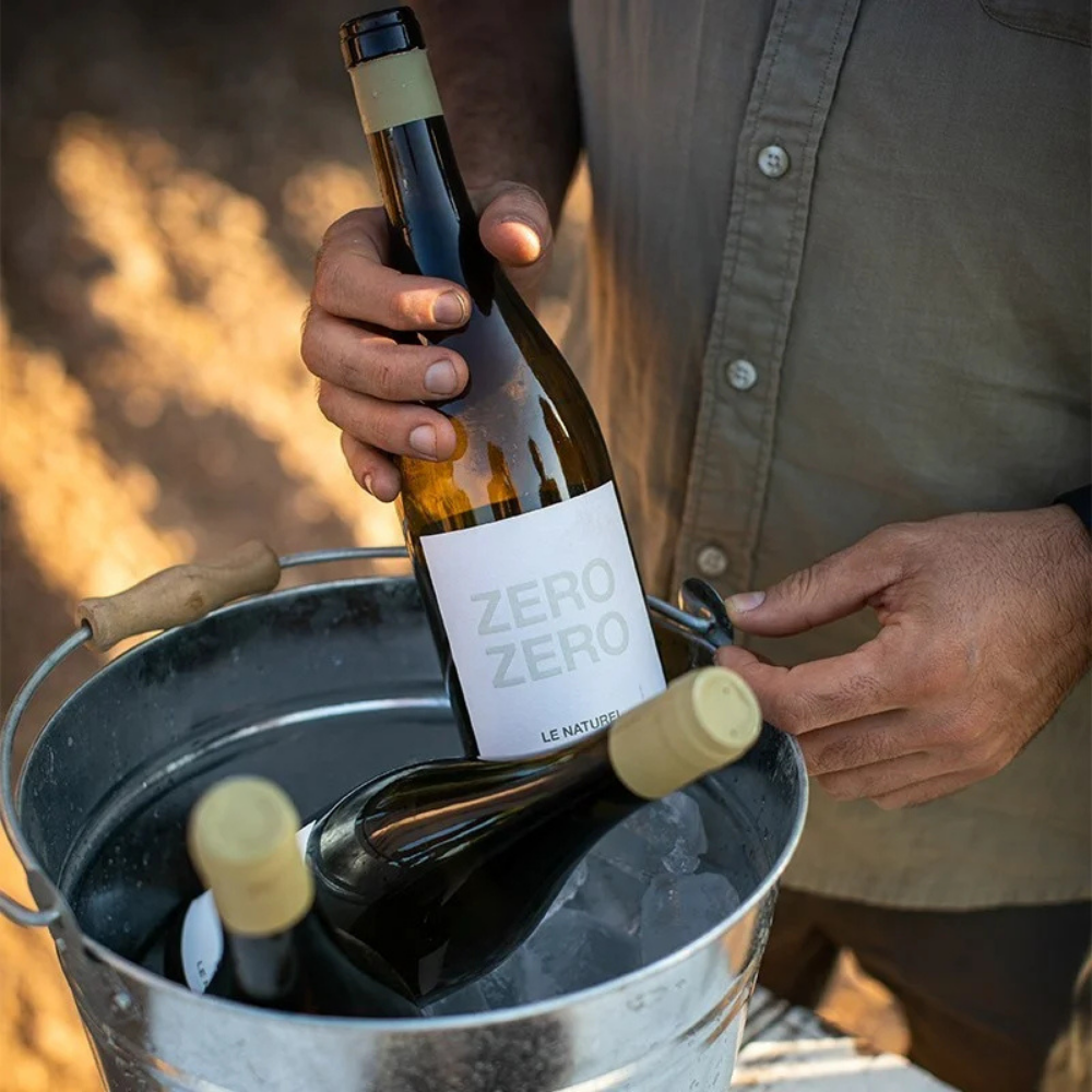 A person holds a bottle of Zero Zero wine above a metal ice bucket containing other wine bottles outdoors.