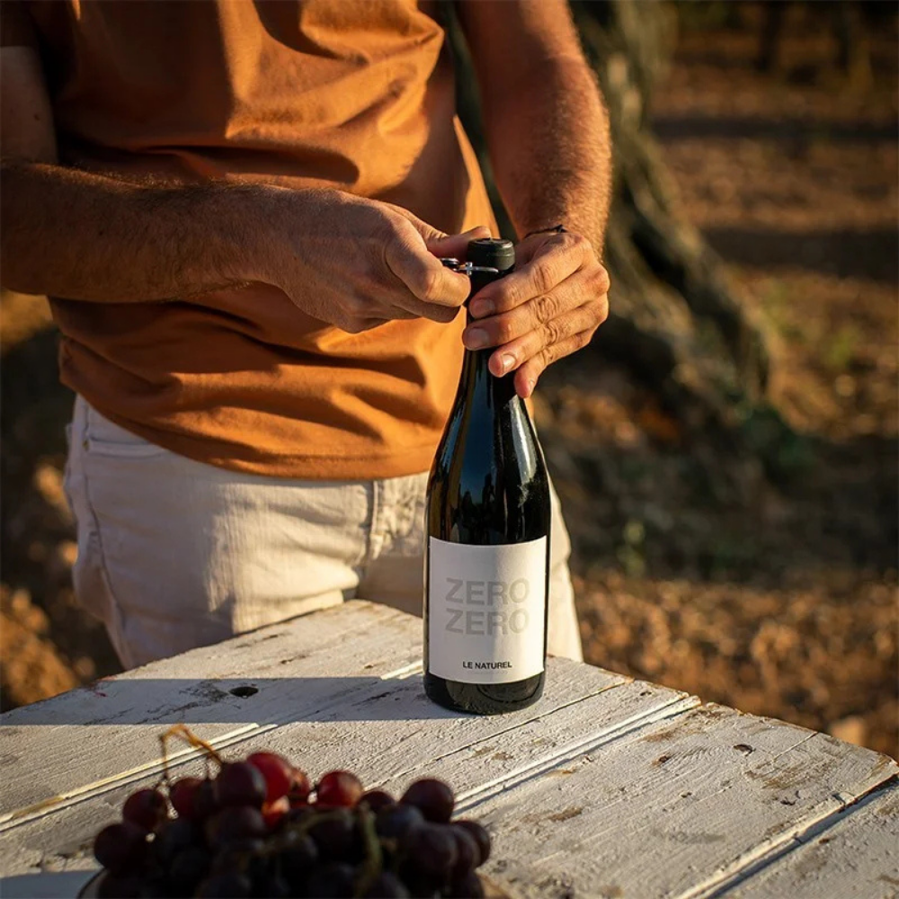 A person in a rust-orange t-shirt and white shorts opens a bottle of Le Moulins Zero Zero sparkling wine on a rustic white wooden table, with a bunch of dark grapes in the foreground and a warm, sunny outdoor background.