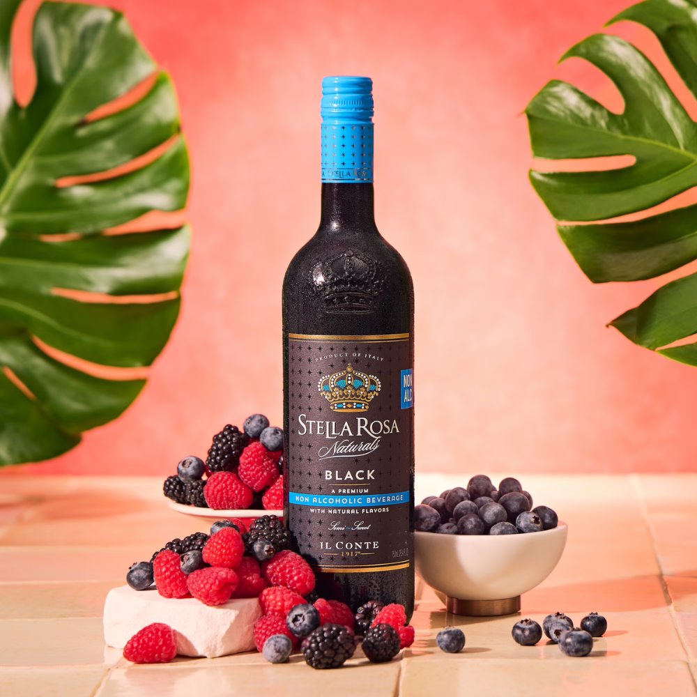A bottle of Stella Rosa Natural Black Blue Raspberry Syrup centered on a peach background, surrounded by fresh raspberries, blueberries, and a small bowl on a tiled surface with large green leaves framing the scene.