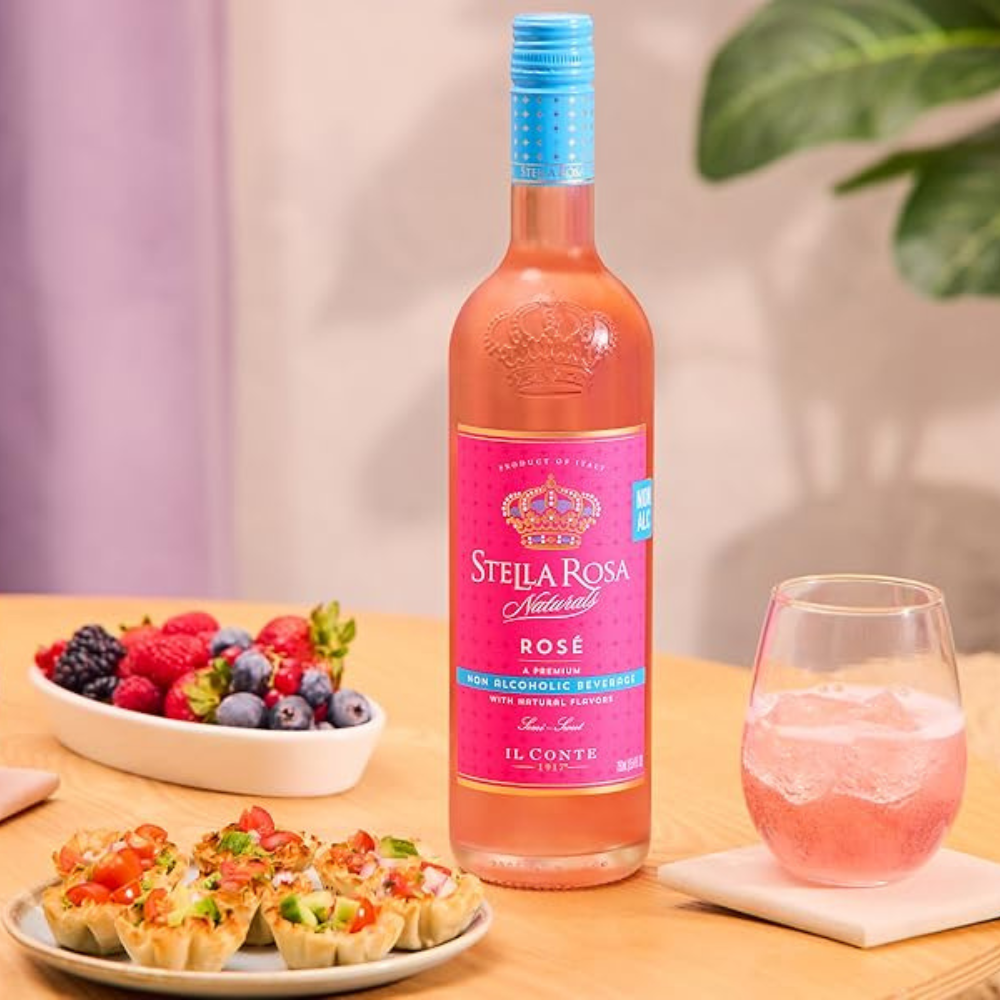A bottle of Stella Rosa Natural Rosé non-alcoholic beverage with a pink label and blue cap, set on a wooden table next to a glass of pink drink, a bowl of fresh berries, and small appetizers.